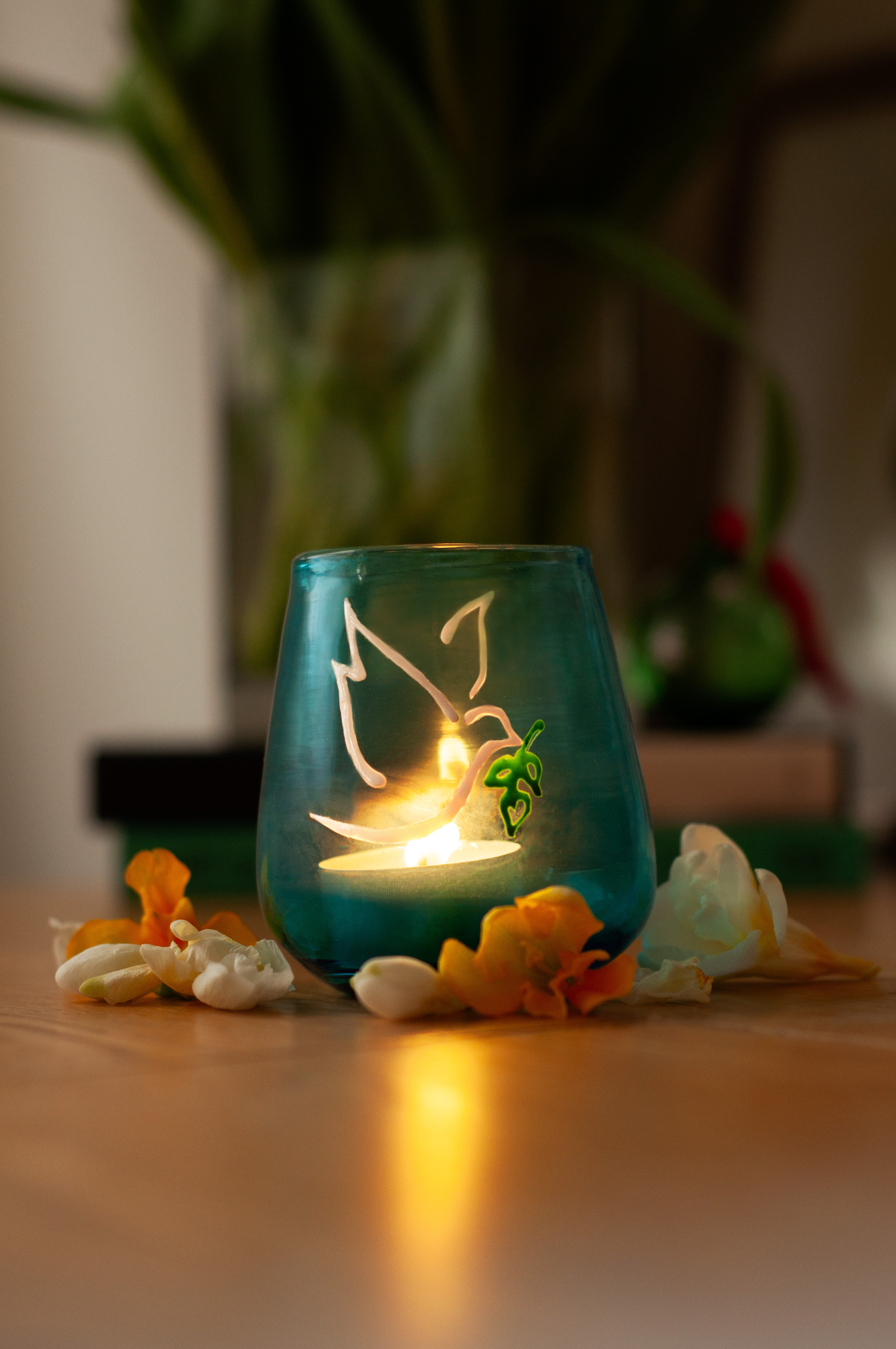 A hand-blown turquoise glass Peace Light with a lit candle inside with painted symbols of a dove and olive branch on the glass surface. Made in Bethlehem